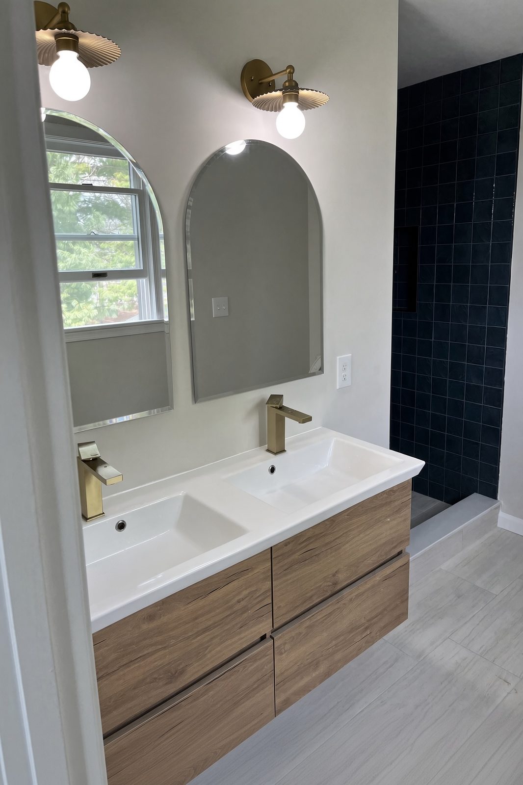 Double oak vanity with gold faucets, arched mirrors, and a dark tile shower beyond