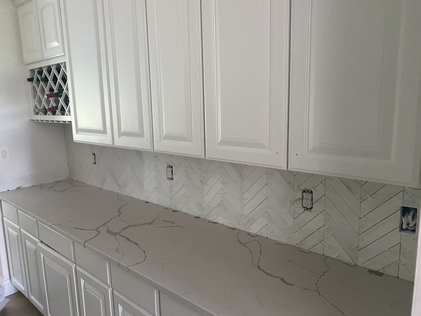 White kitchen with a marble-look herringbone backsplash, quartz counters, and a wine rack
